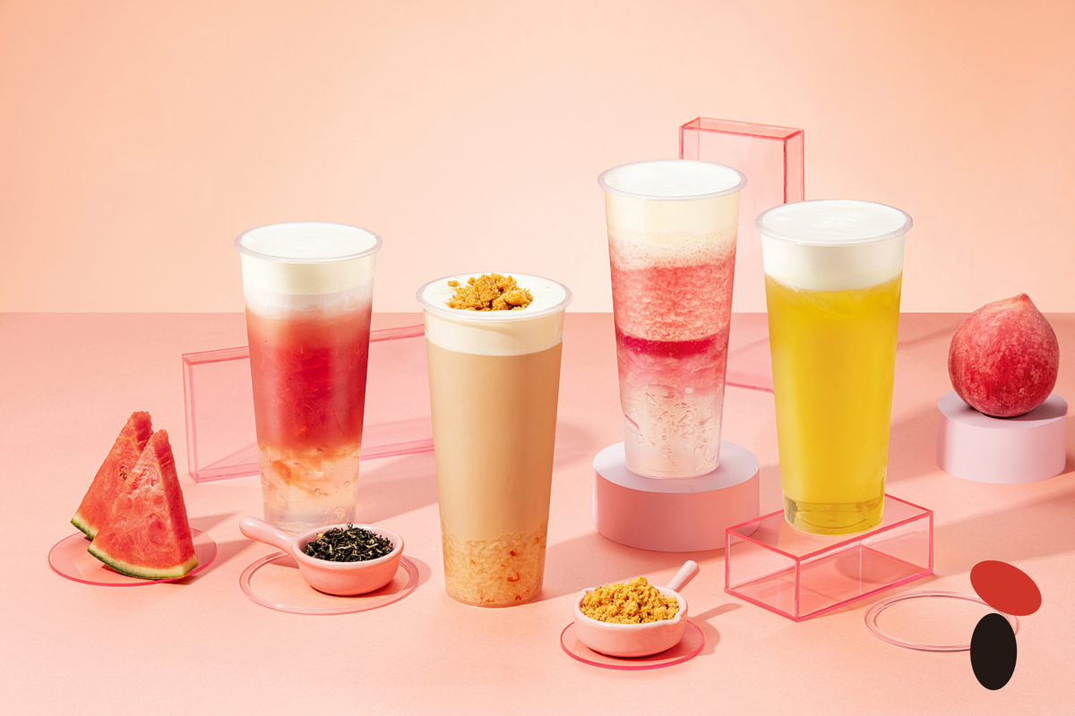 Four bubble tea drinks in different colours beside sliced fruit and small bowls of toppings on a pink background